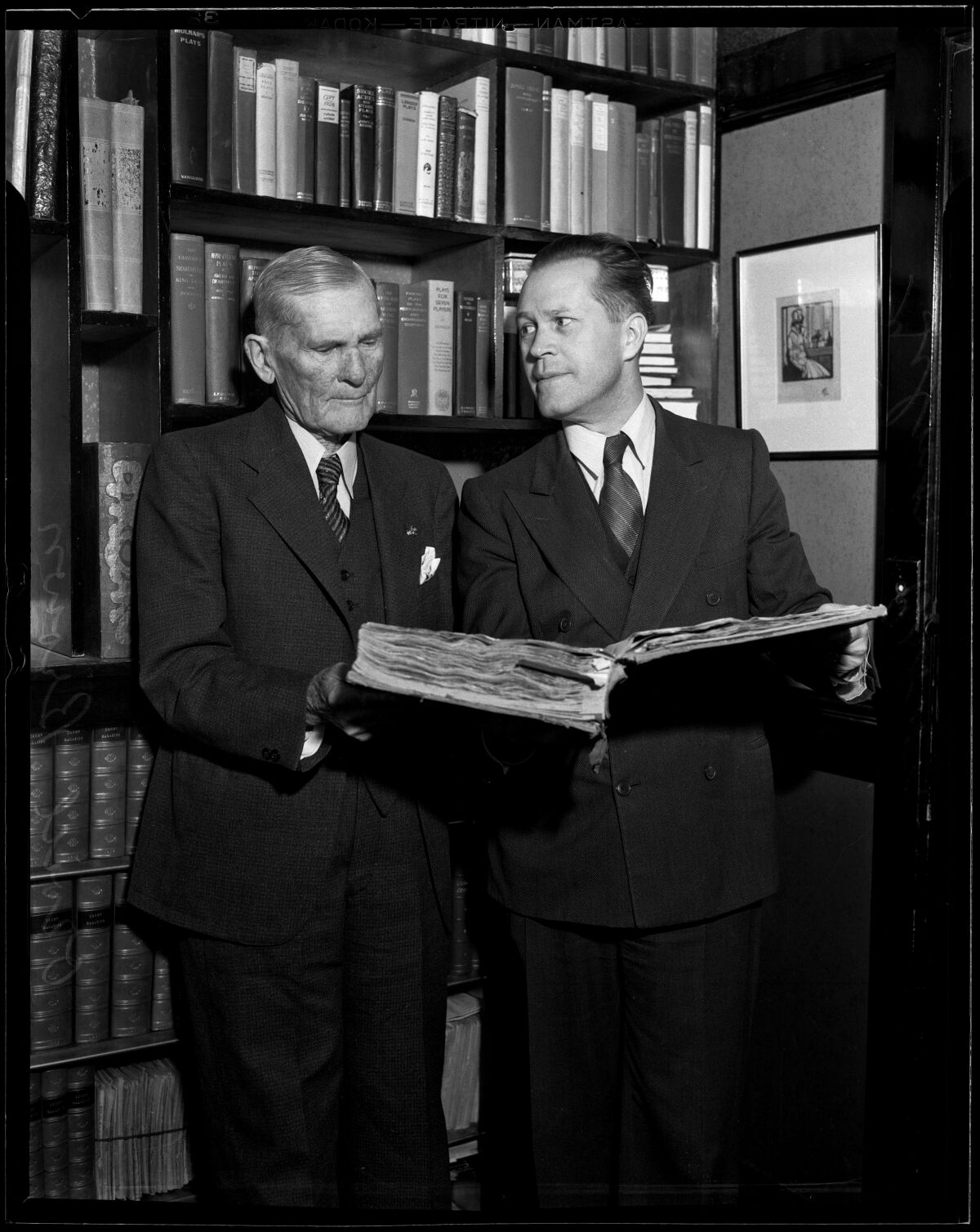 In a black-and-white photo, two men stand in front of a bookcase, one holding a large open book. 