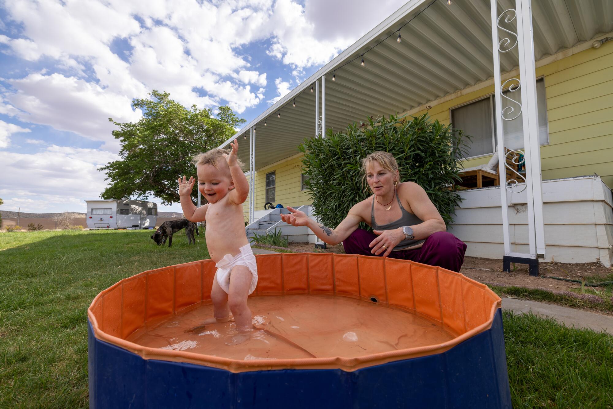 一名幼儿在室外婴儿泳池里戏水，他的母亲在一旁观看。  