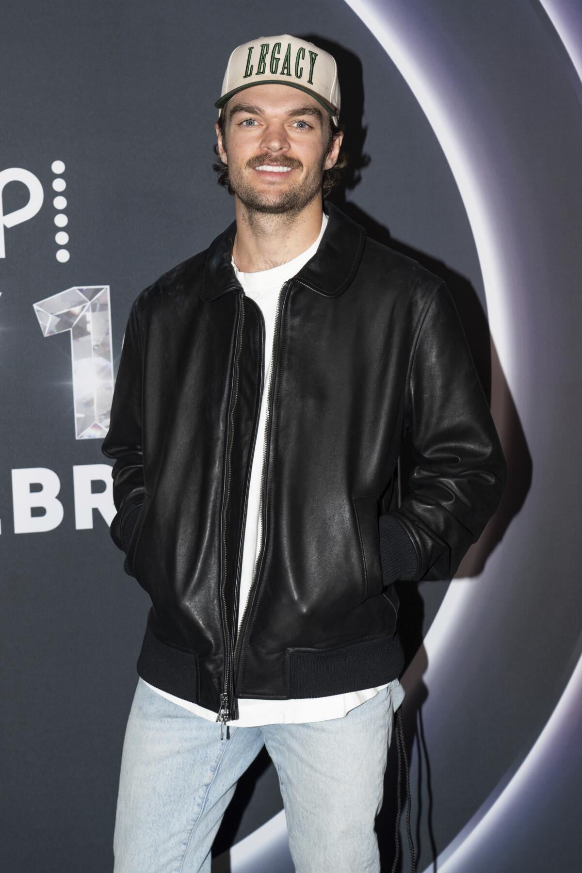 Conner Smith smiles in jeans and a trucker cap that reads "Legacy" with his hands in the pockets of his shiny black jacket