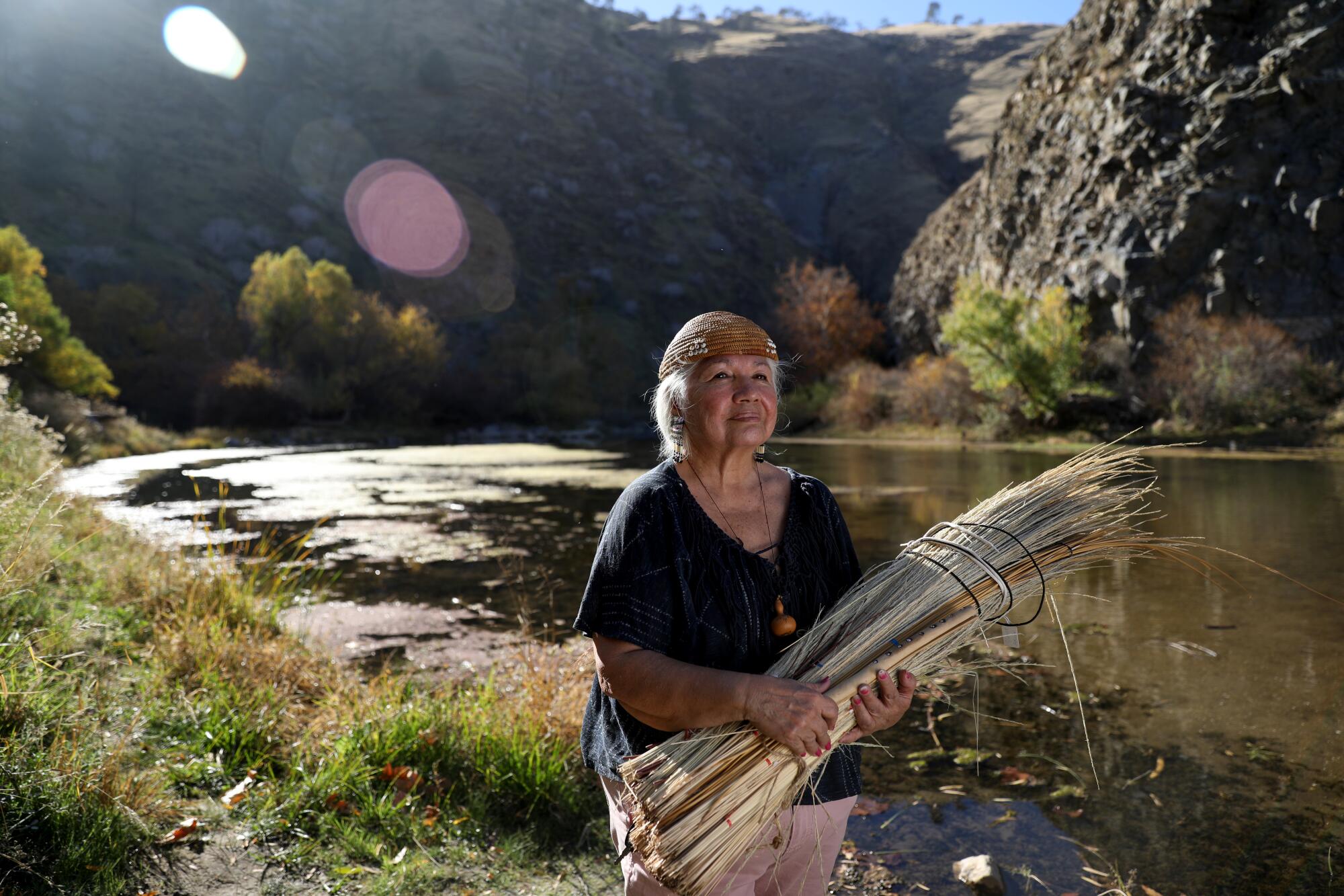 A woman stands beside a river holding reeds.