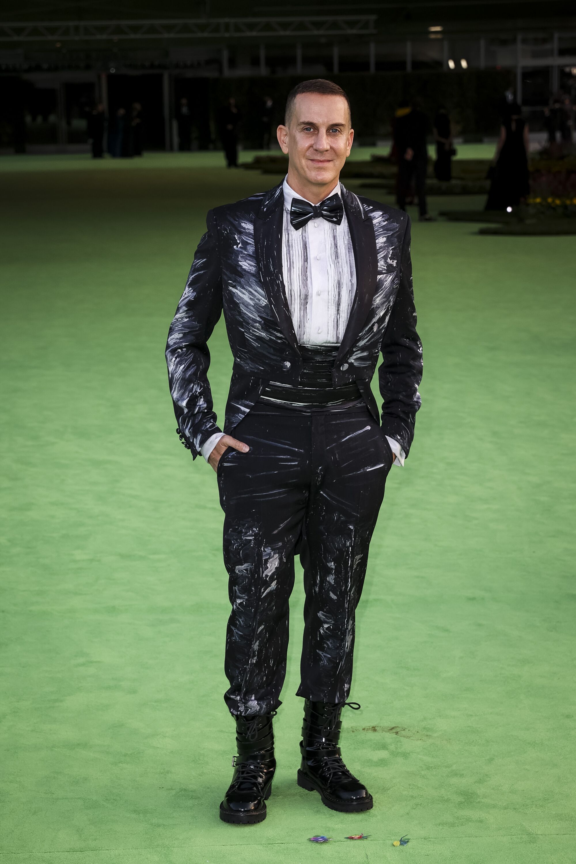A man in a patterned black tuxedo and bow tie posing on a green carpet