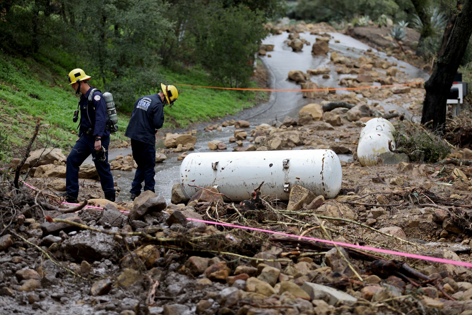 Two people look at rocks and debris, including two white tanks, blocking a road.