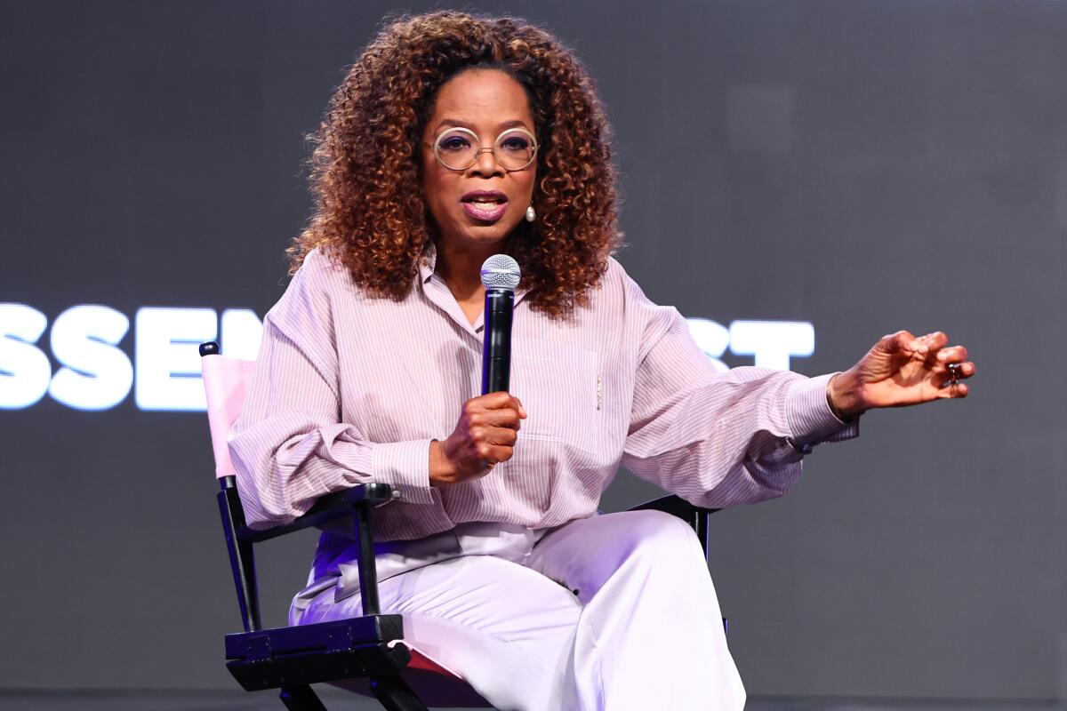 Oprah Winfrey sits in a director's chair onstage talking into a microphone and gesturing with one hand