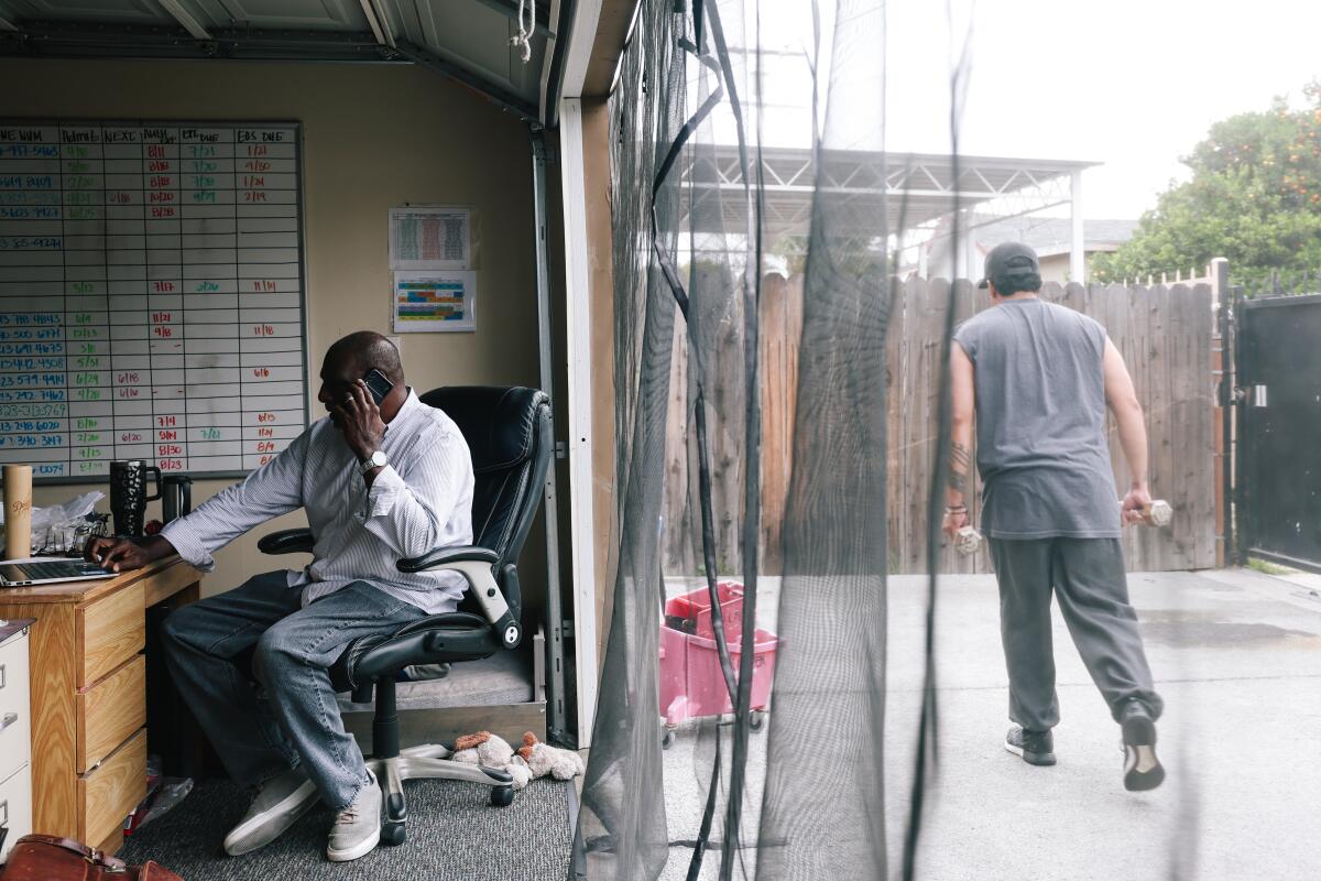 Kalain Hadley, left, speaks on his cellphone while seated at a desk as a person exercises nearby beyond mesh netting