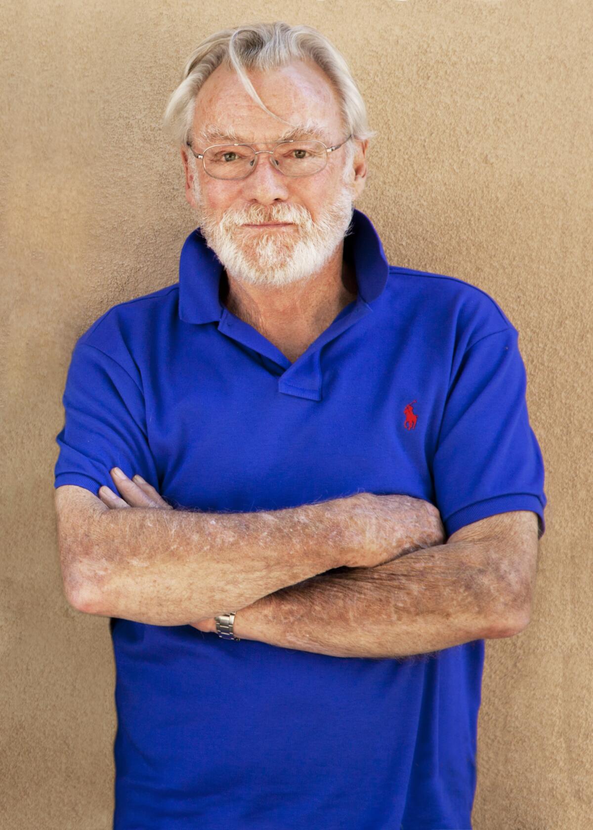 John Morris wears a blue polo with an orange insignia as he poses for photos in front of a beige wall