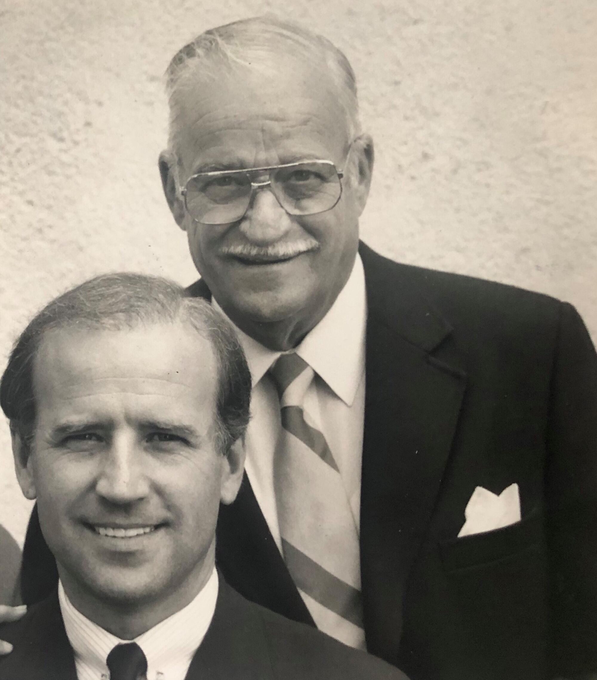 A man in a suit and tie with another older man, both wearing a suit and tie and smiling
