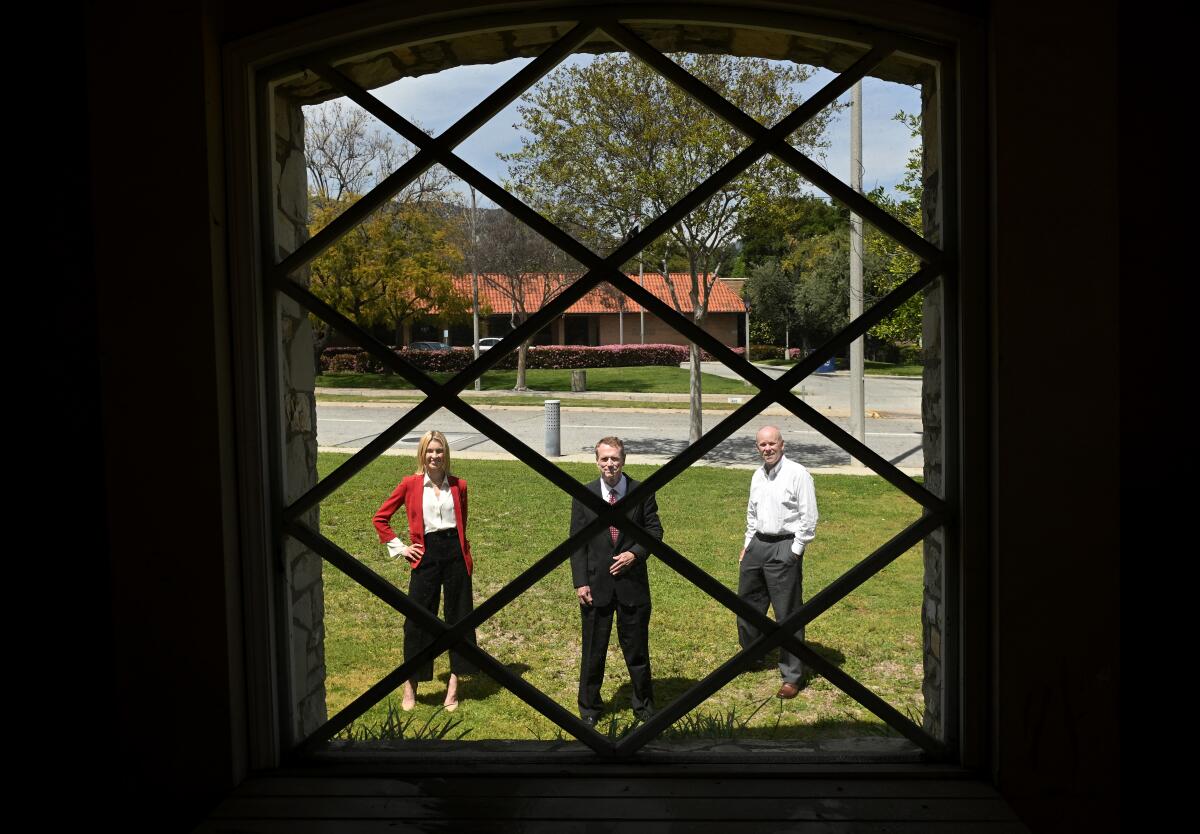 A woman and 2 men stand on a lawn, seen from inside through a window with grilles in a horizontal diamond-shaped pattern