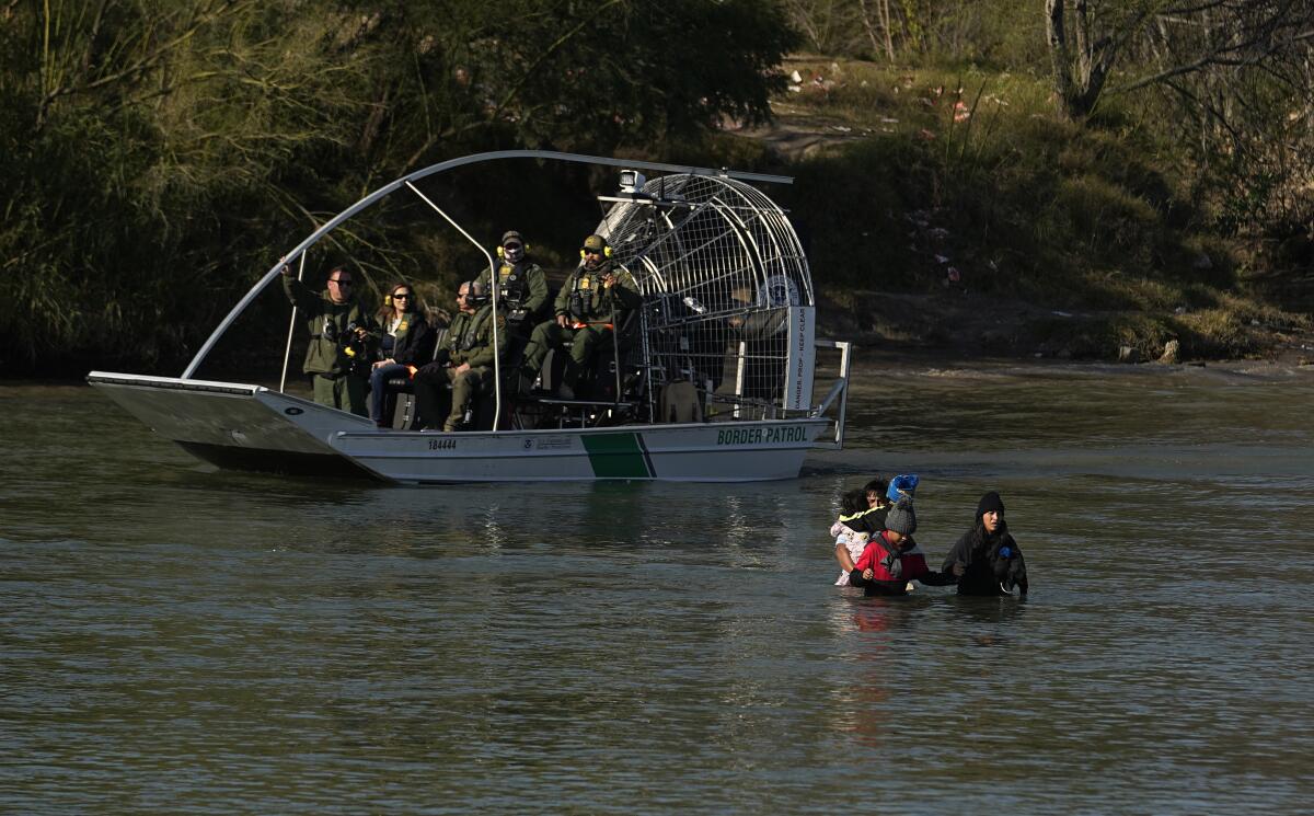 Border agents in fan boats watch as people wade across a river 