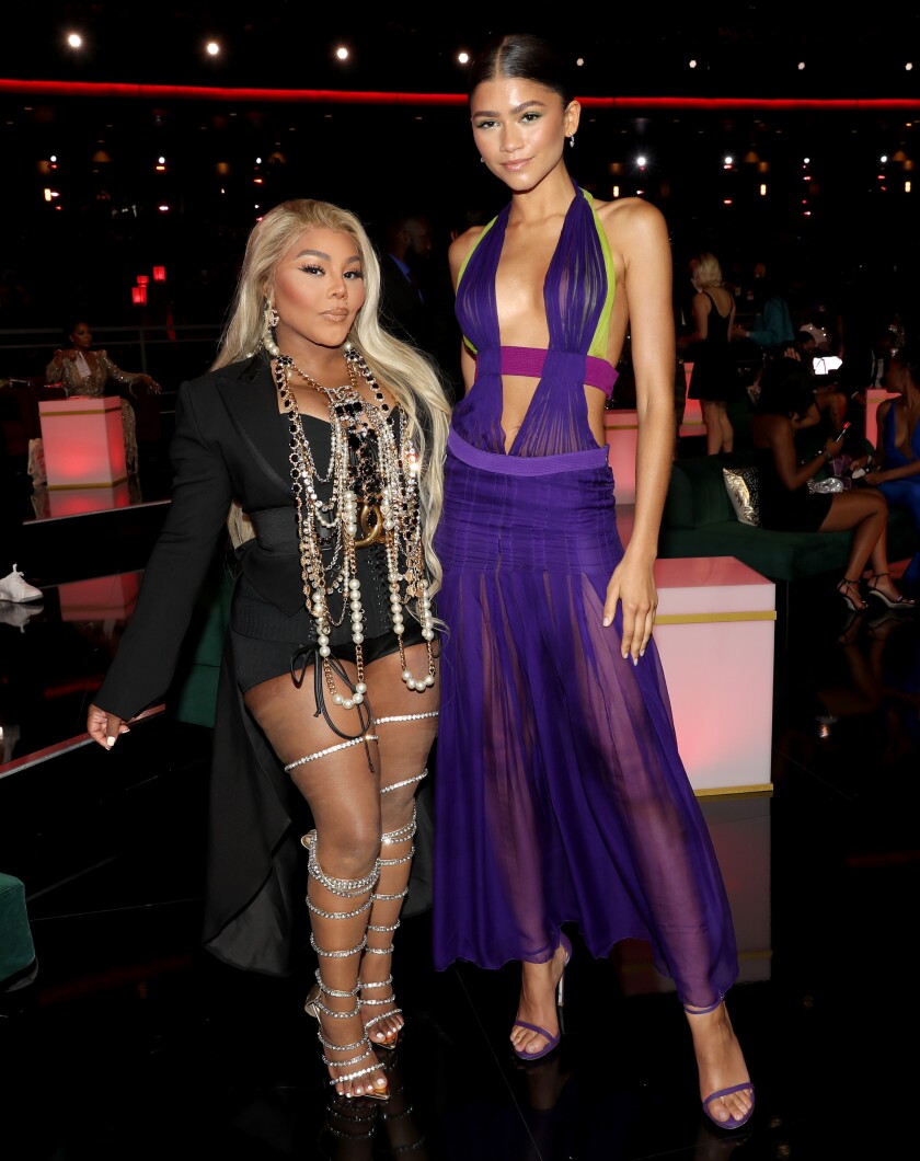 Two women in formal wear on a red carpet.  One is wearing a black dress with a necklace, the other is in a transparent purple dress