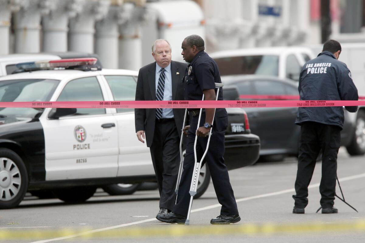 A man in dark clothes, using crutches, walks near another man, in suit and tie, standing near a patrol car