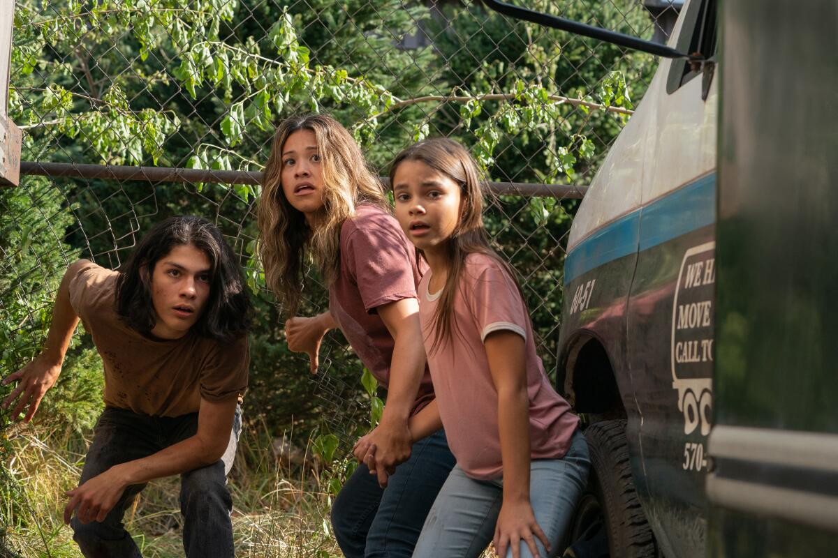 A woman and a young man and a young girl crouch in front of foliage and beside a truck. 