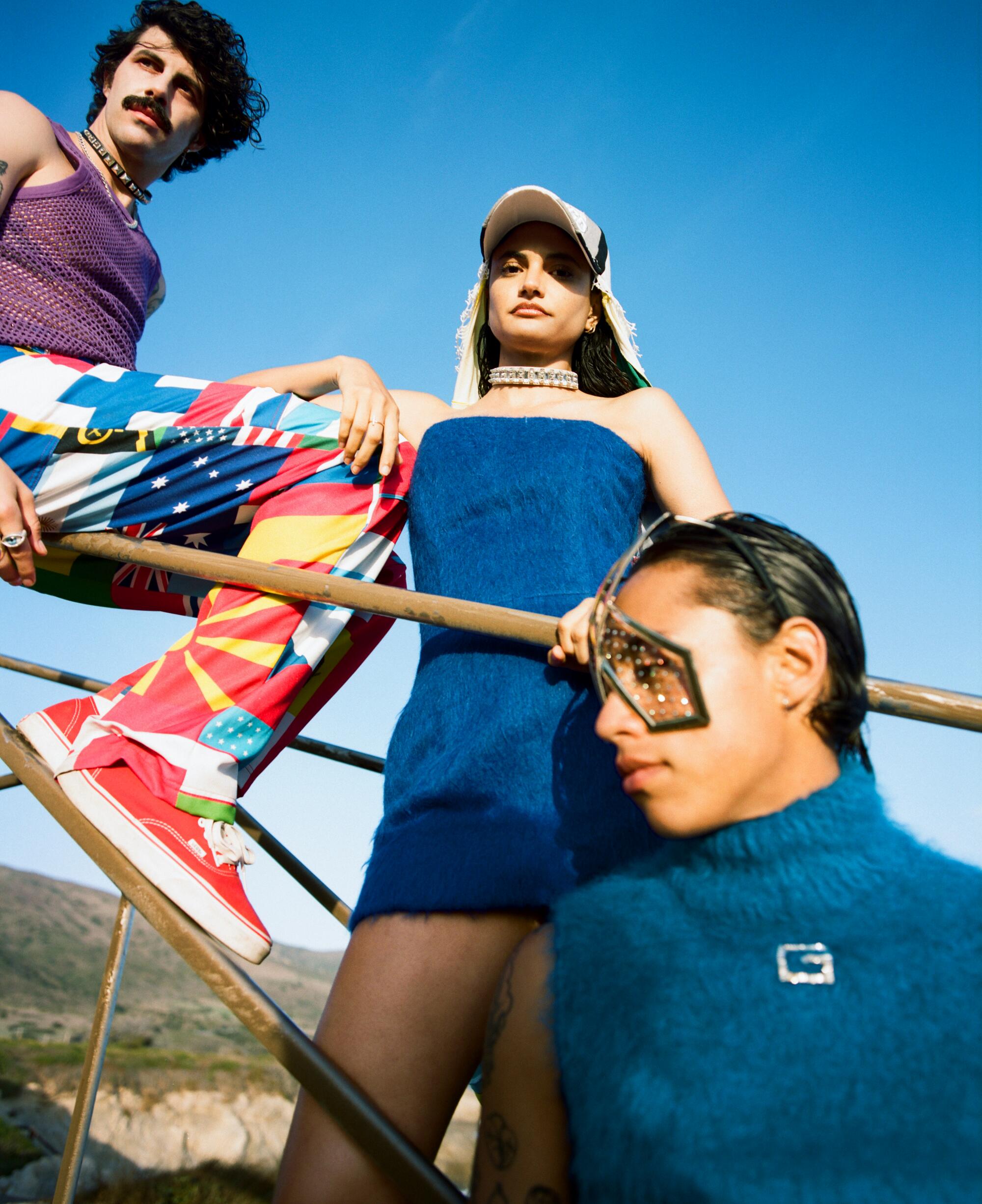 Three people wearing colorful outfits hang out by a railing on the beach.