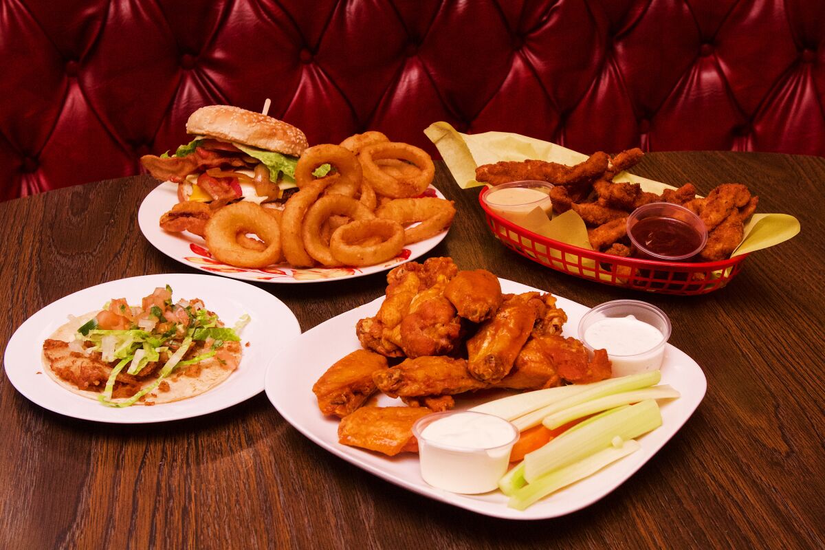 A fish taco, bacon cheeseburger, basket of chicken tenders and plate of wings on a table in front of a red booth. 