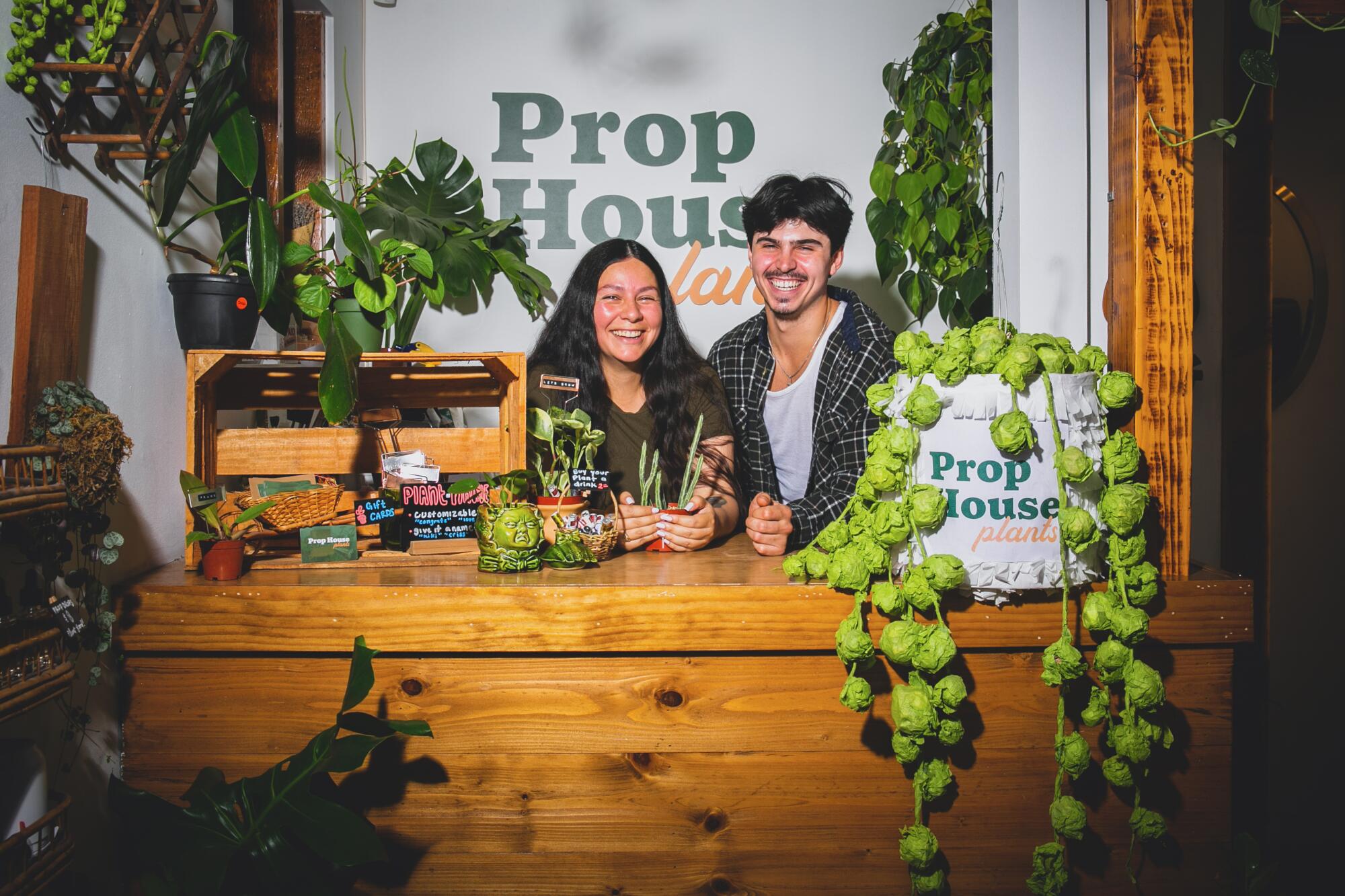 A young woman and a young man stand behind a counter smiling, with plants around them.