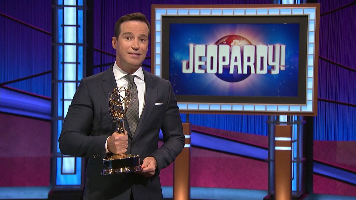Mike Richards in a suit holding an Emmy trophy on the 'Jeopardy!' set