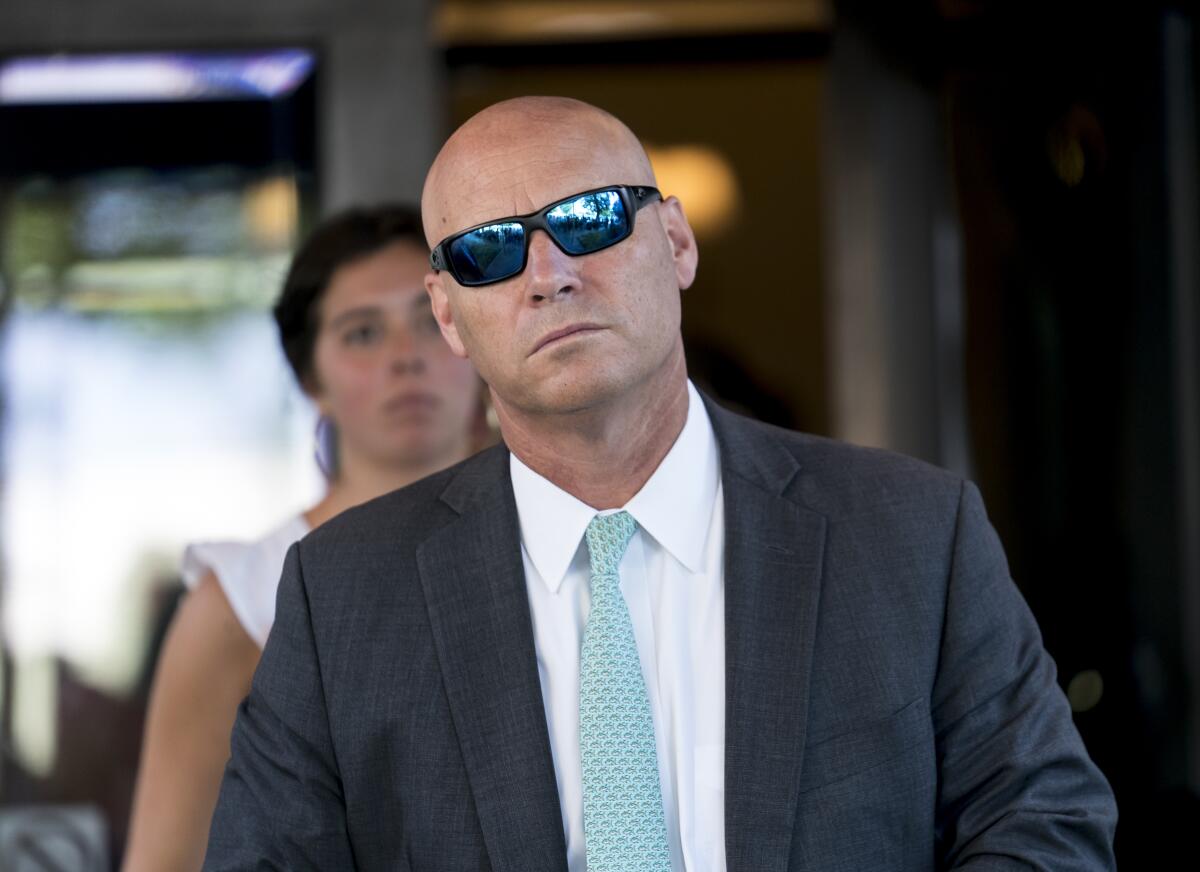 A man with a clean-shaven head, wearing a dark suit, pale green tie and sunglasses 