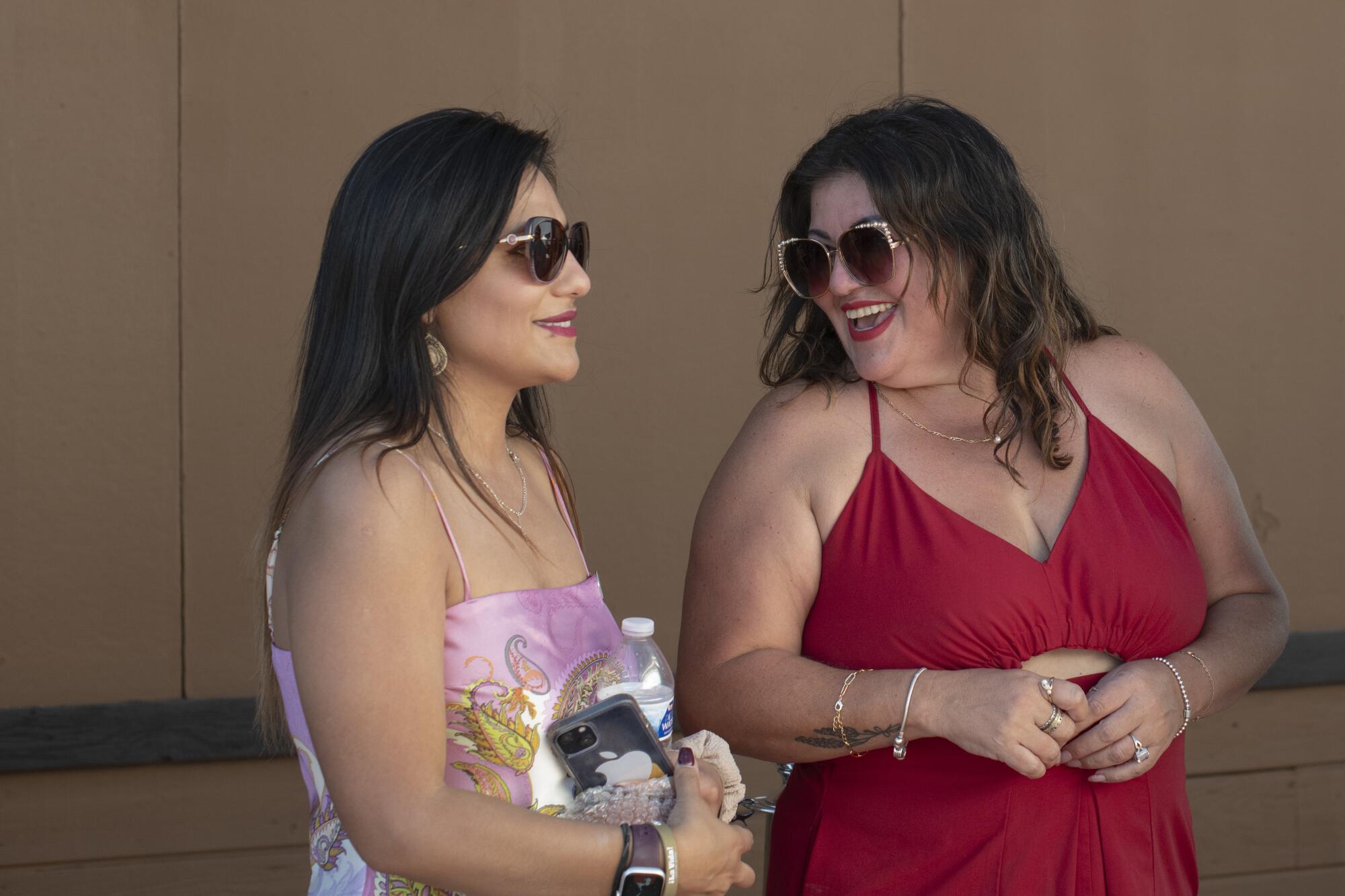 Two smiling women with dark hair and sunglasses standing together, one in a flowery pink dress, left, and the other in red 