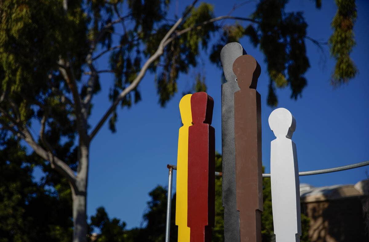 A sculpture shows five stylized figures of varying heights in different colors against a blue sky beneath a tree.