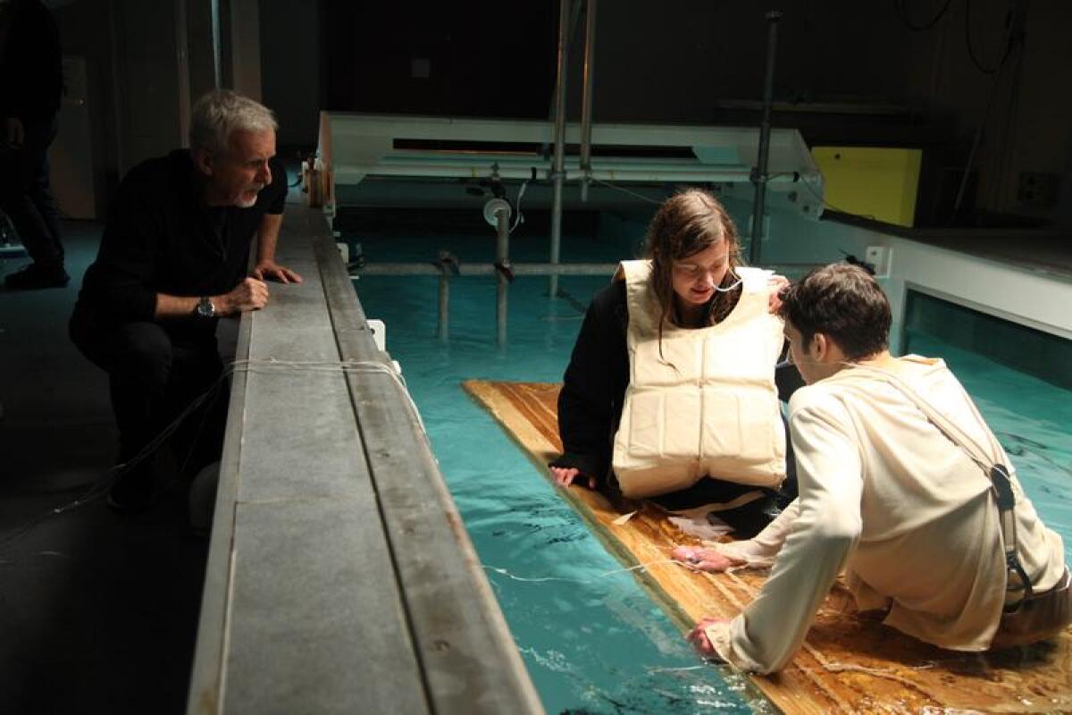 A man bends over a pool where two stunt people sit on a floating door.