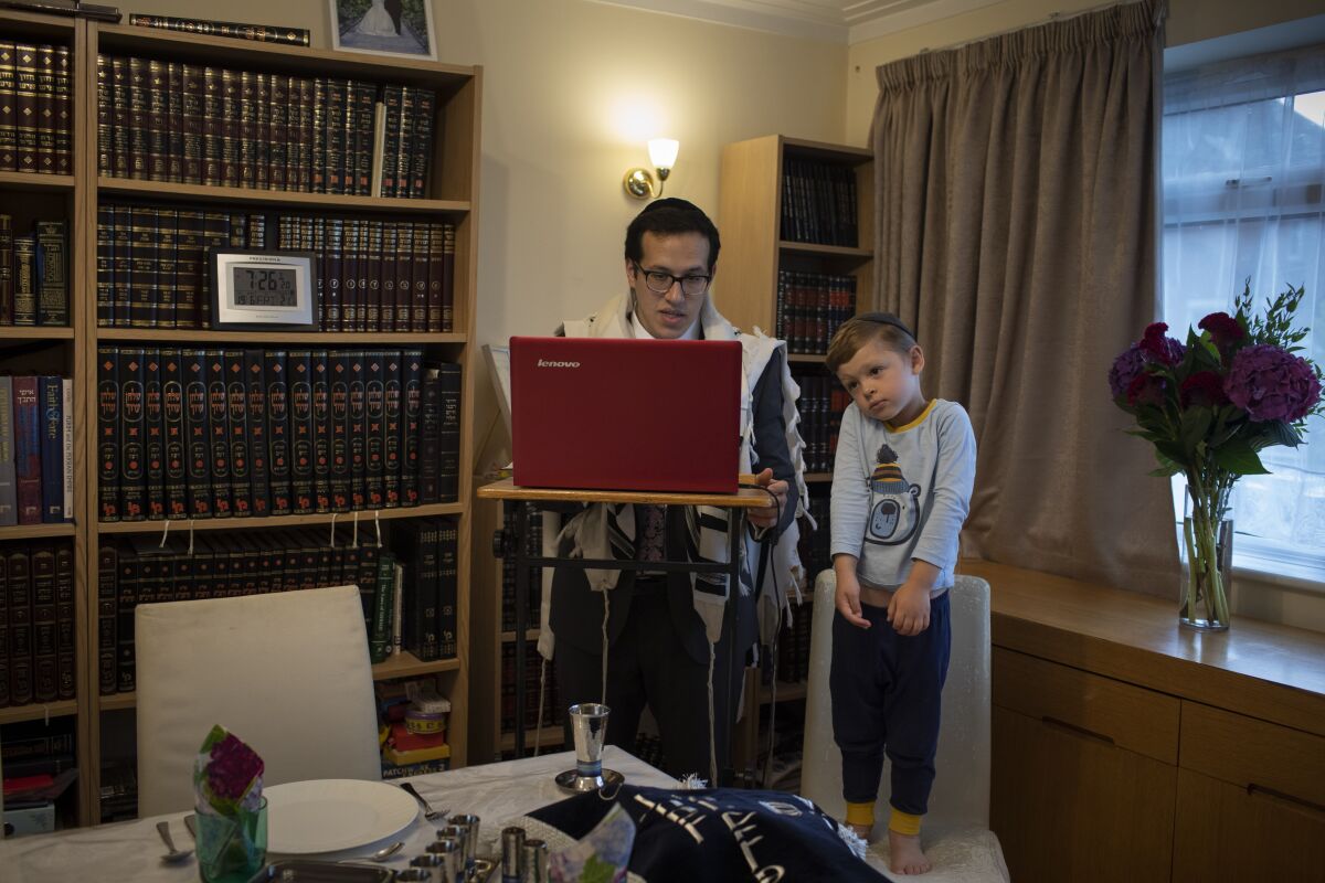 As his 3-year-old son Tzvi looks on, Rabbi Mordechai Chalk leads a service for his congregation via a teleconference app from his home in London on Friday, June 19, 2020, just before sunset. Taking services online-only has been particularly challenging for the Orthodox Jewish community, members of which are proscribed from using electronics on Shabbat, their day of rest. (AP Photo/Elizabeth Dalziel)