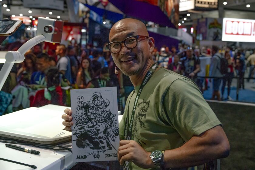A bald man wearing glasses and smiling while holding up a drawing in front of a crowd