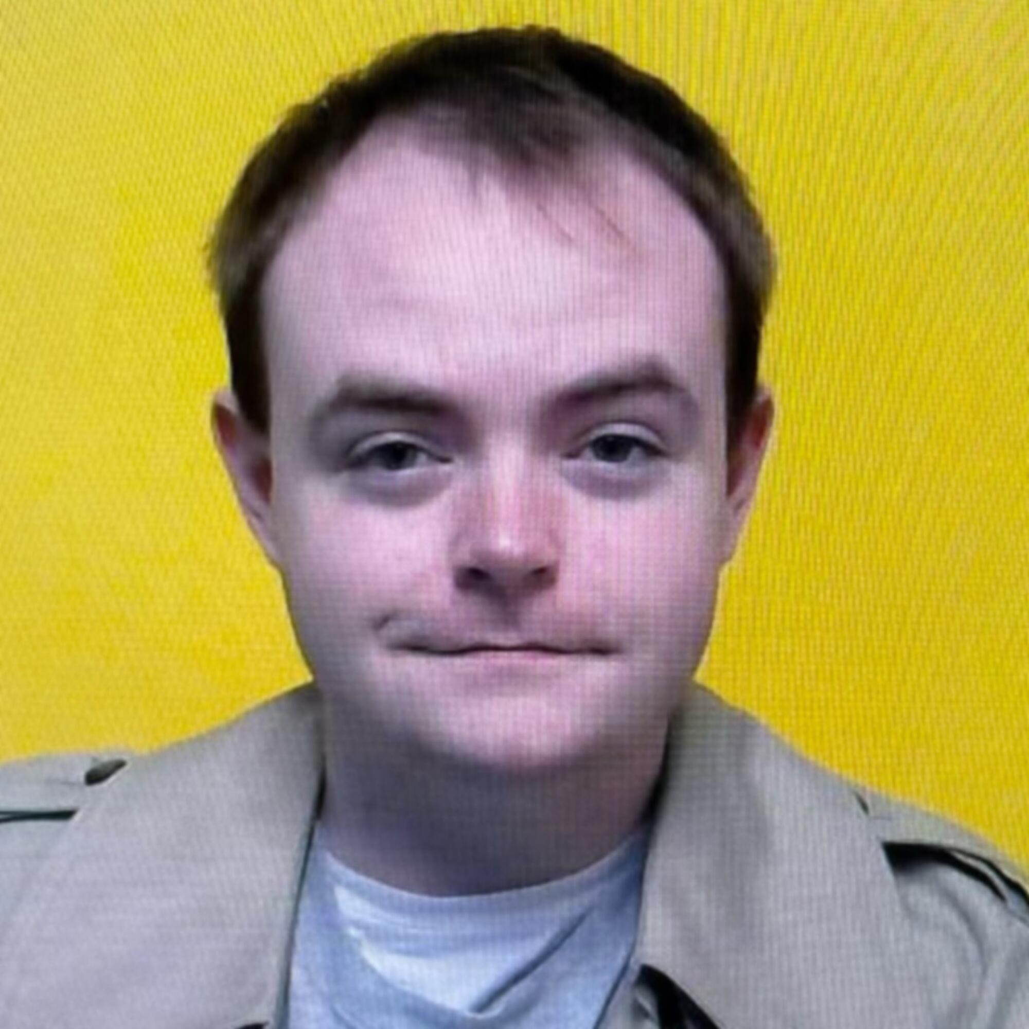 A head-and-shoulders frame of a young man in a tan coat slightly smiling, looking at the camera, against a yellow background.