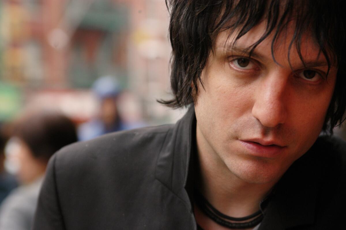 Jesse Malin is posing for a closeup photo wearing a gray top with his bangs flowing down his face.