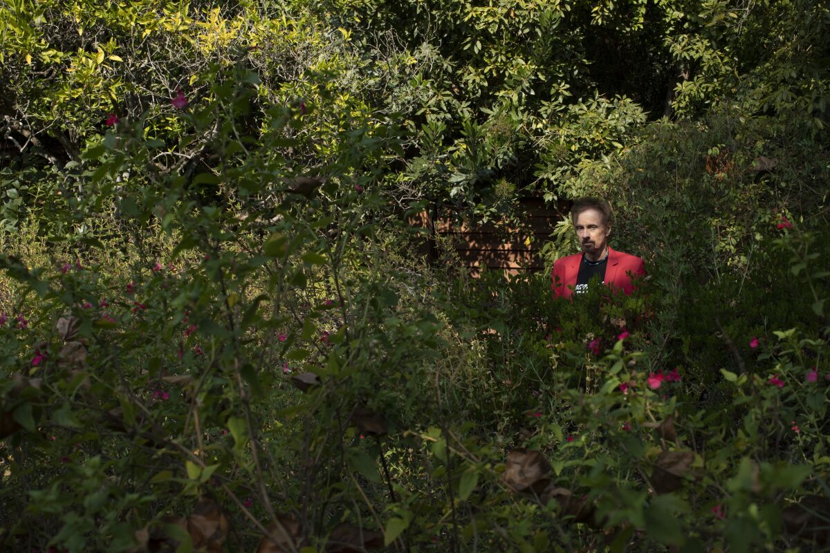 Author T.C. Boyle, wearing a black T-shirt and red jacket, stands amid dense shrubbery.