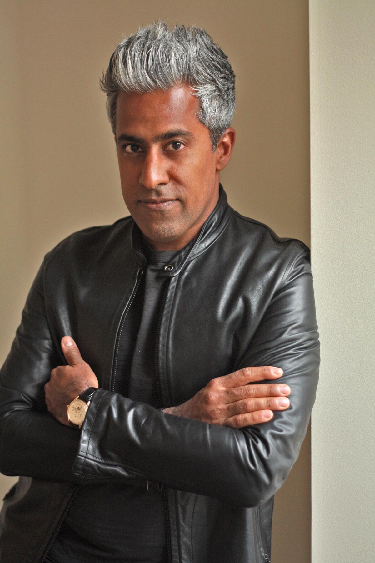 A man with gray hair and a leather jacket poses against a wall, his arms folded.