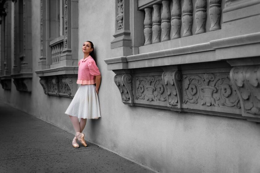 A ballerina dressed in a 1950s-style costume stands against an old building.