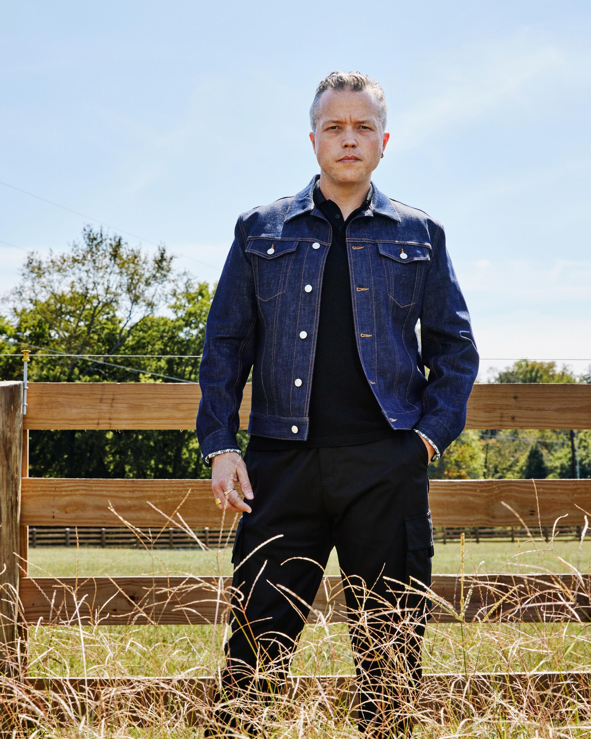 A country-rock singer-songwriter in a denim jacket stands outside in front of a wood fence