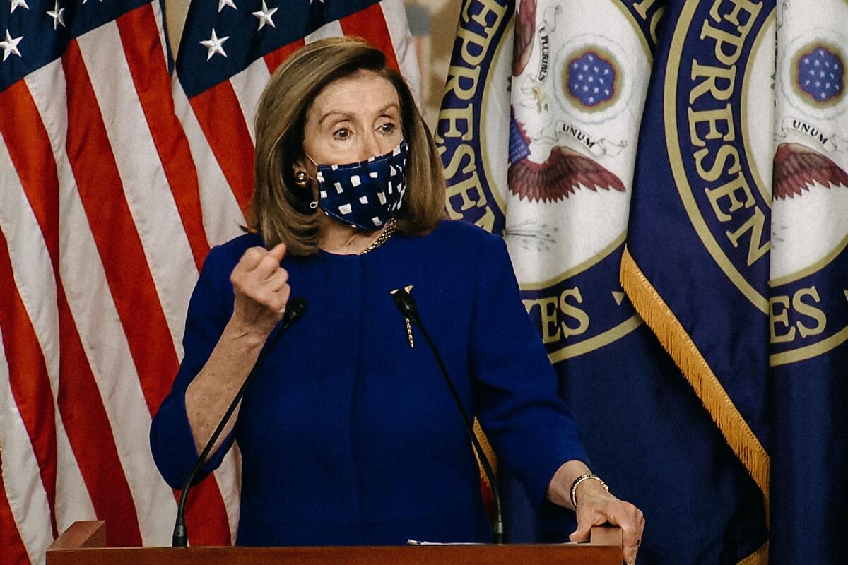 Nancy Pelosi, wearing a cloth mask, raises a clenched hand as she speaks, with several House and U.S. flags behind her.