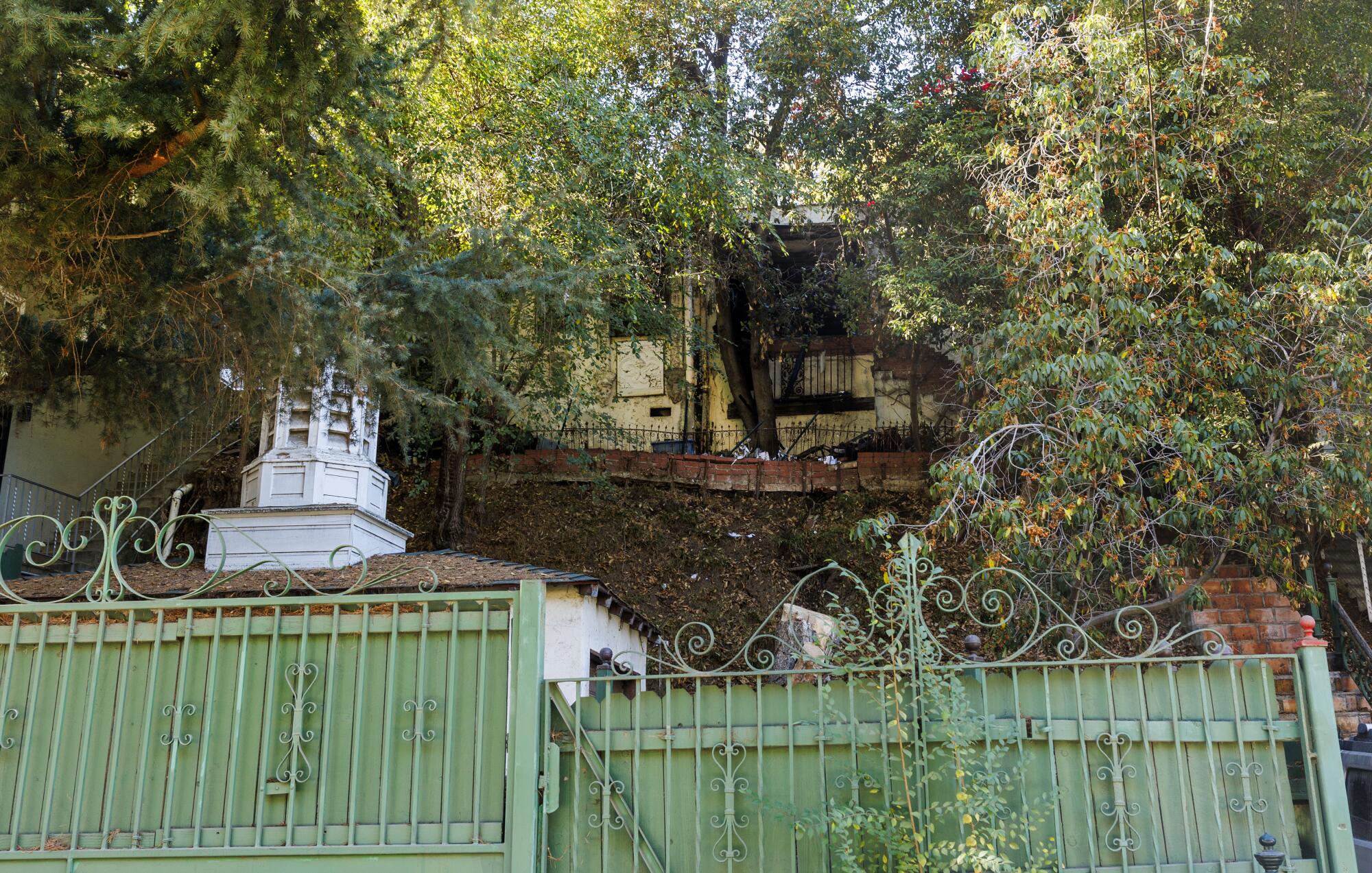 A yellow house with fire damage on a hillside above trees, a small white building and a green metal fence in the foreground