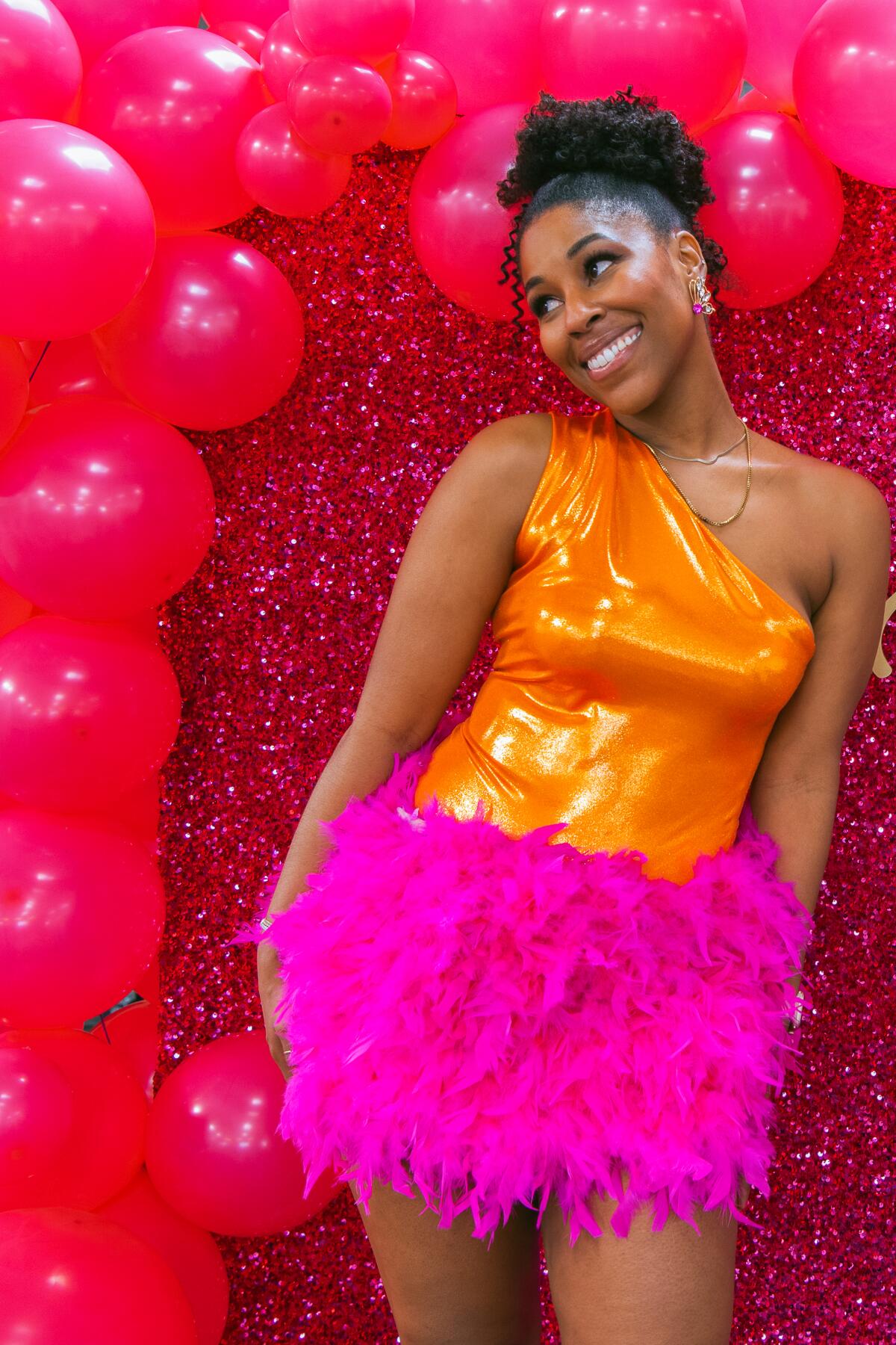 A portrait of Aryn Morris, wearing an orange top and feathered pink skirt, against a pink background