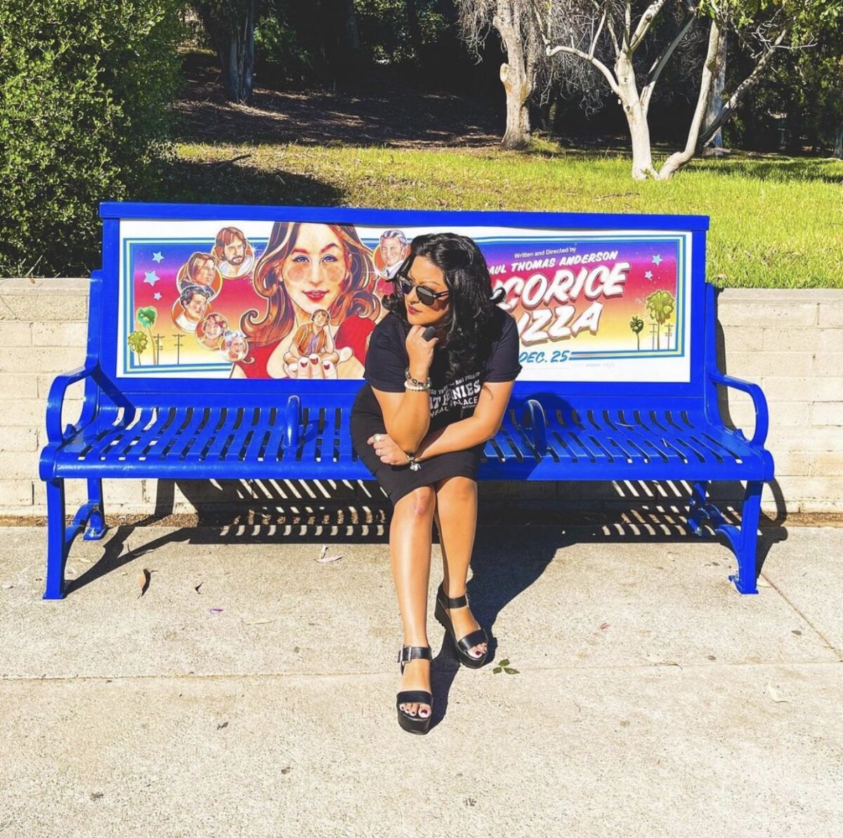A woman sits on a bench with a movie poster behind her.  