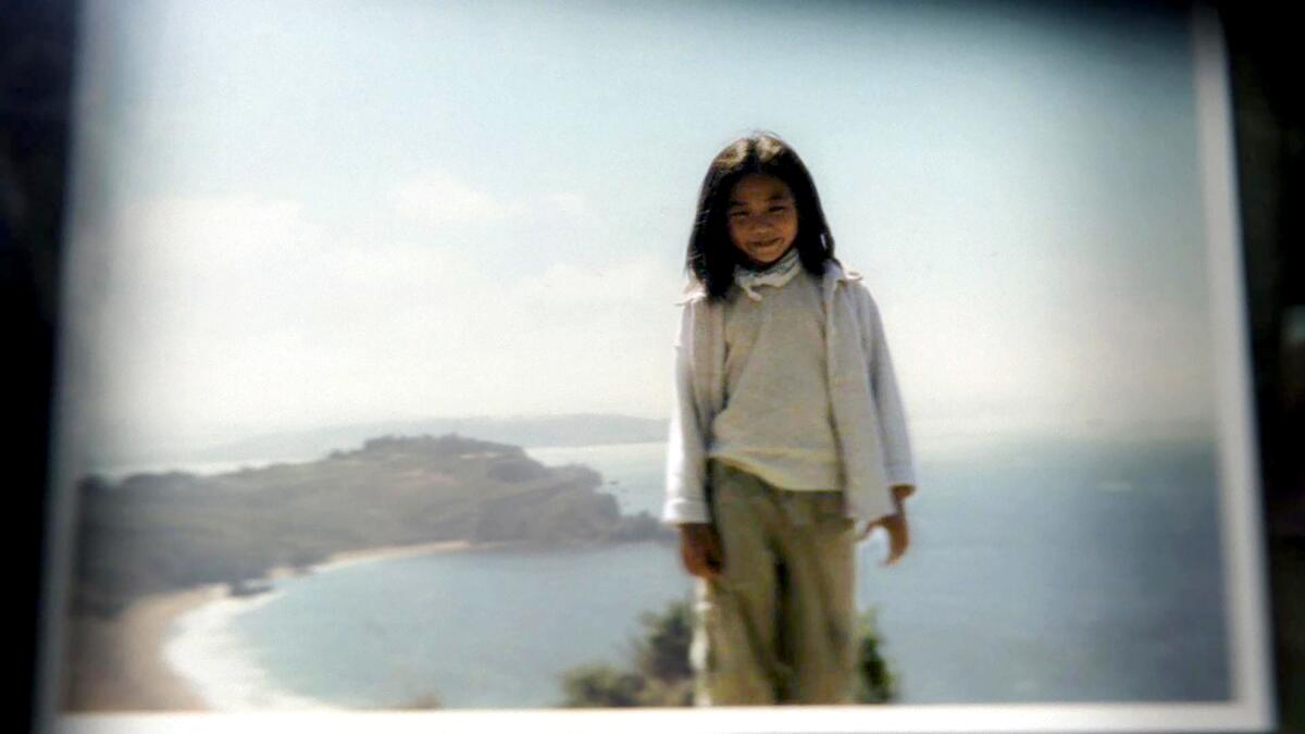 A photo of Rachel Lee as a child in a white jacket, shirt and khaki pants standing in front of a cove with blue water.