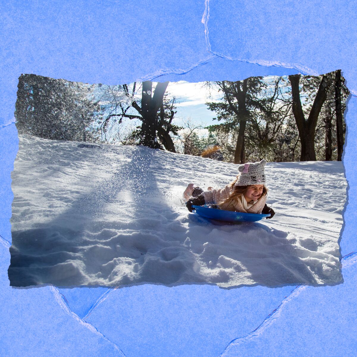 A young girl in winter coat, hat and gloves smiles while sledding on a snowy hill.