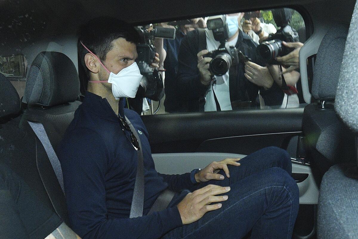 A masked man with dark hair and dark clothes is seated in the back of a vehicle with photographers outside his window