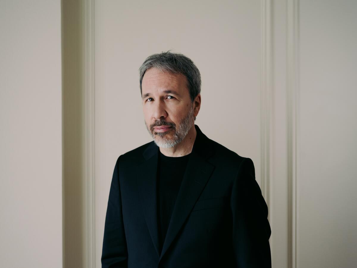 Denis Villeneuve poses in a black jack shirt with a serious expression on his bearded face in front of a white background