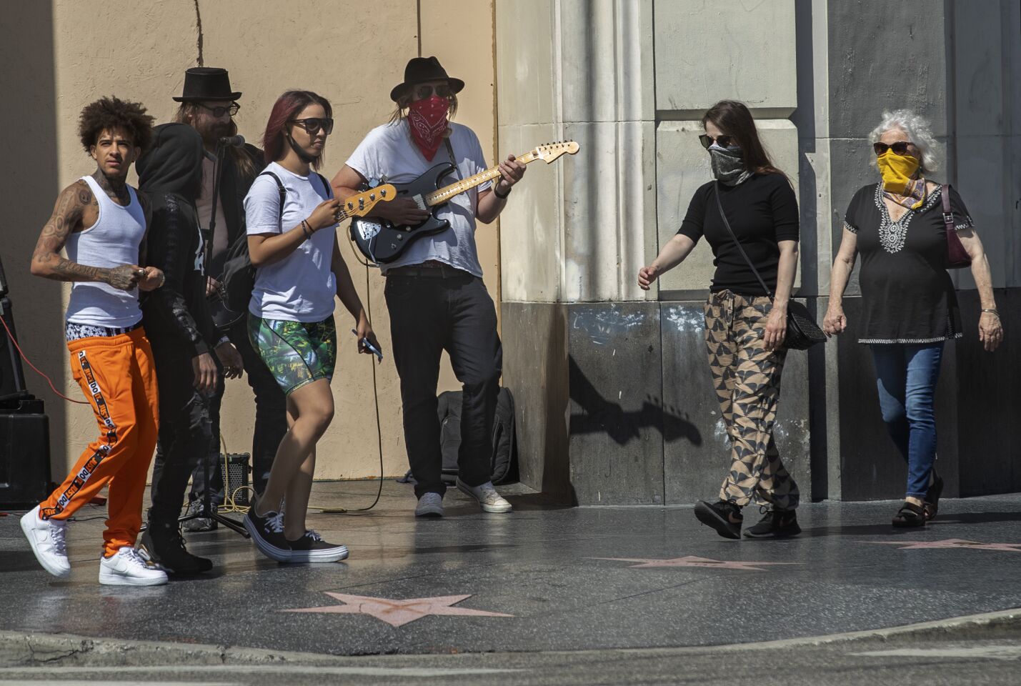 Pedestrians, some with face coverings, some without, walk past musicians at Hollywood Boulevard and Highland Avenue.