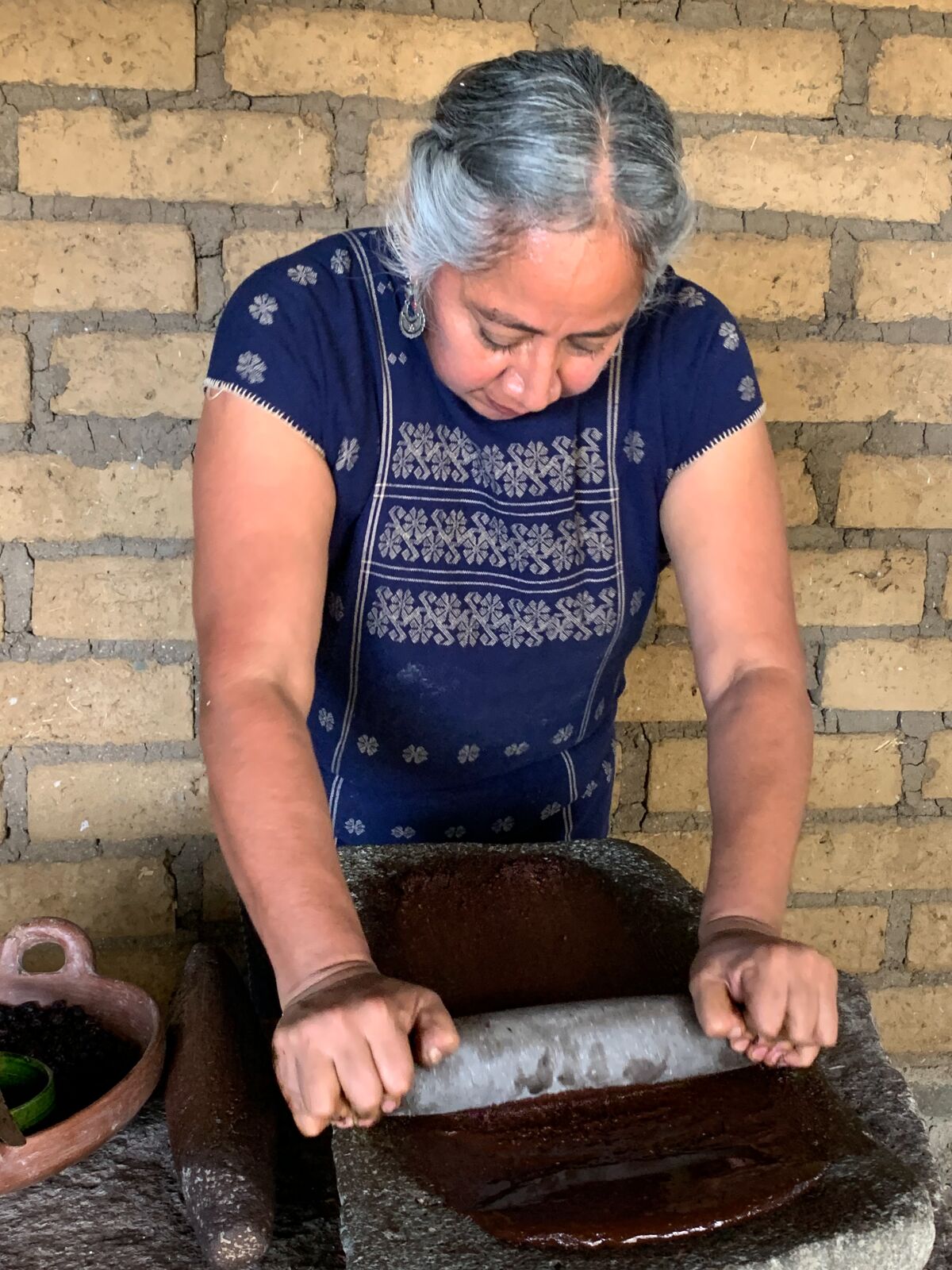 A woman grinds ingredients into a paste on a metate with a stone mano. 