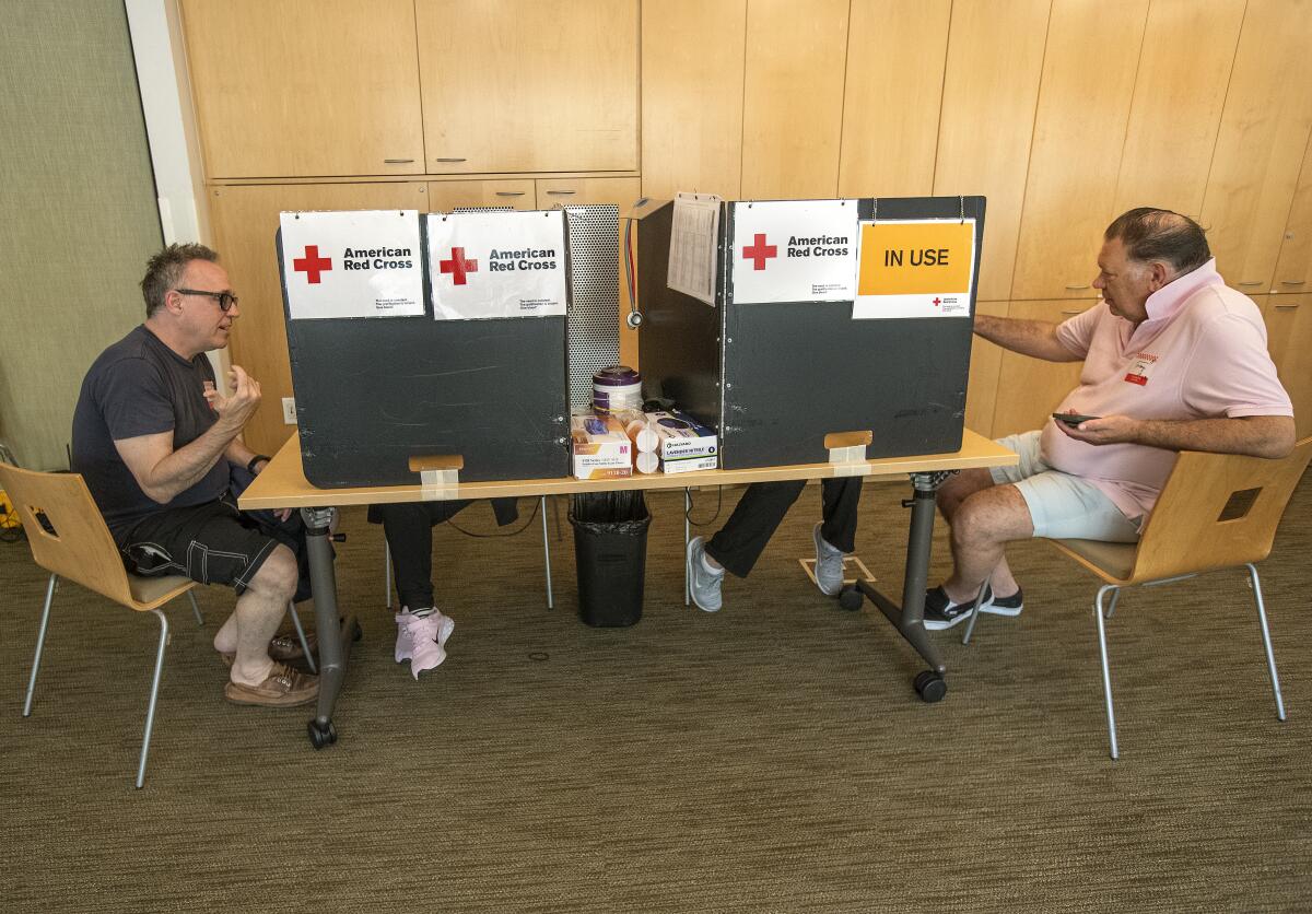 Two men are seated at an opposite side of a table, talking to someone behind privacy shields with "American Red Cross" signs