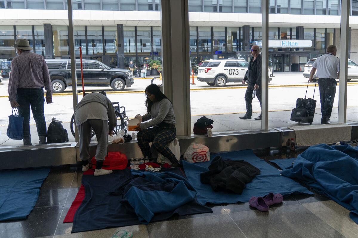 People sit near glass windows, with blue mats lining the floor. Outside the windows are travelers with suitcases and cars