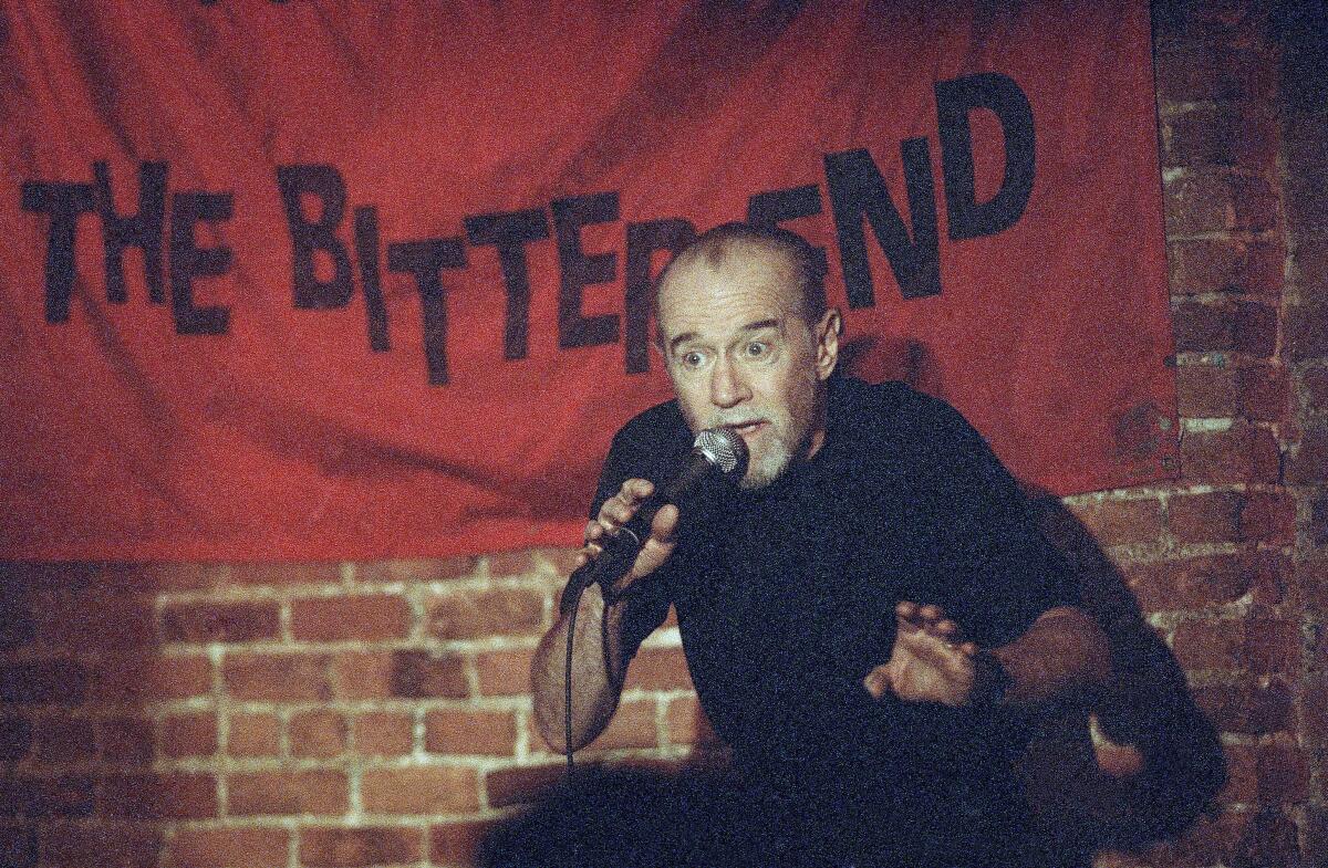 George Carlin wears a black T-shirt and speaks into a mic while standing in front of a brick wall.