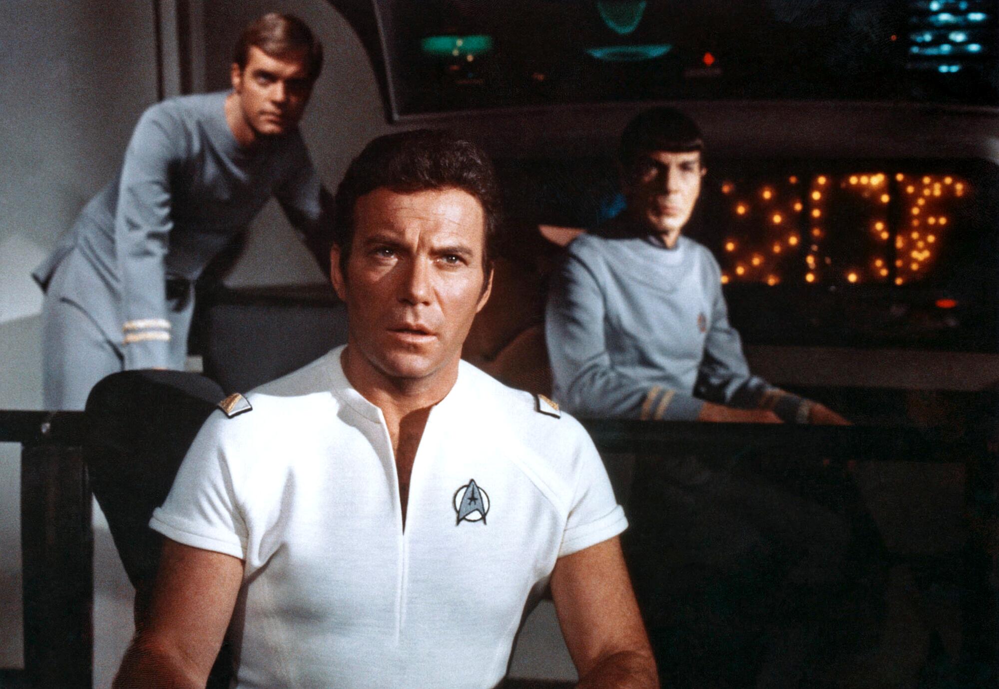 A man in a white uniform sits in the foreground of a spaceship with two men behind him.