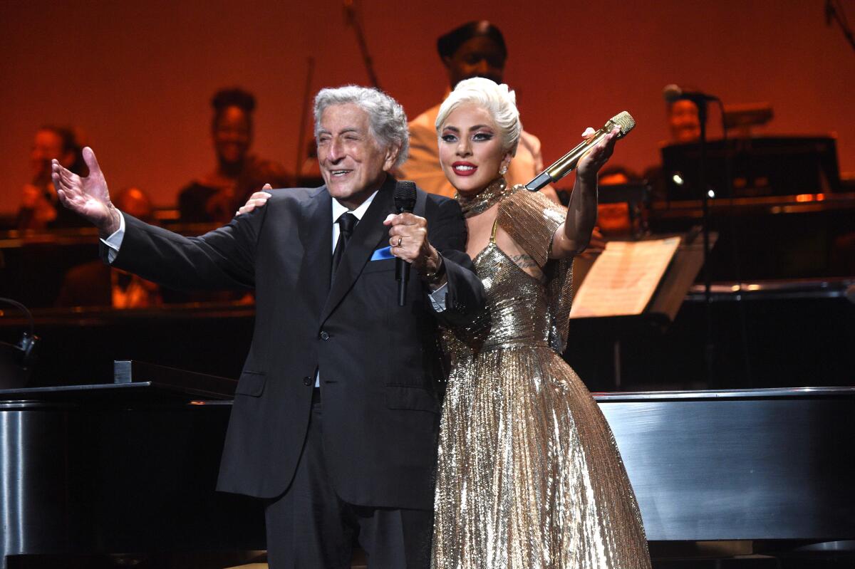 Tony Bennett and Lady Gaga stand on stage with their arms out.