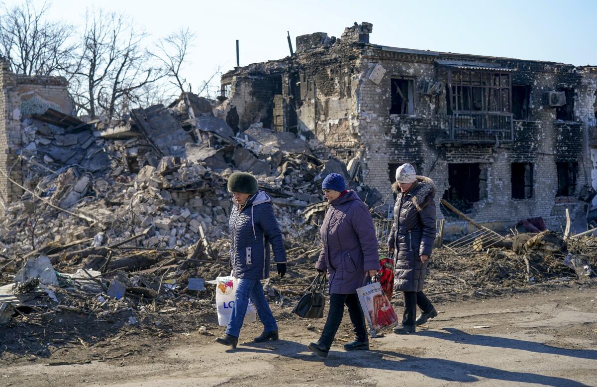 People in coats and hats carry plastic bags past destroyed buildings.