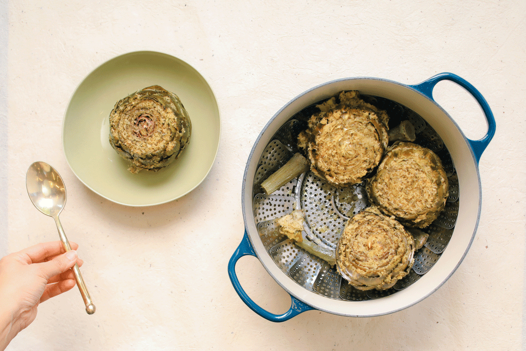 A pot with stuffed artichokes in a steamer basket next to a stuffed artichoke on a plate, with animated spoons moving around