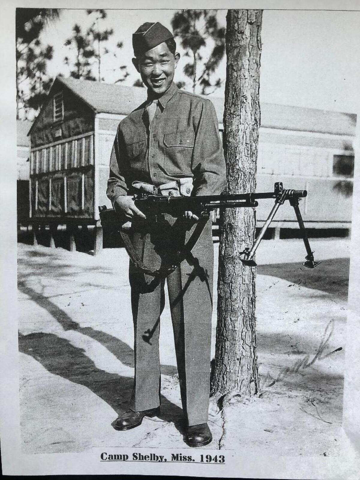 Young Frank Wada stands outside a barracks, holding a gun.