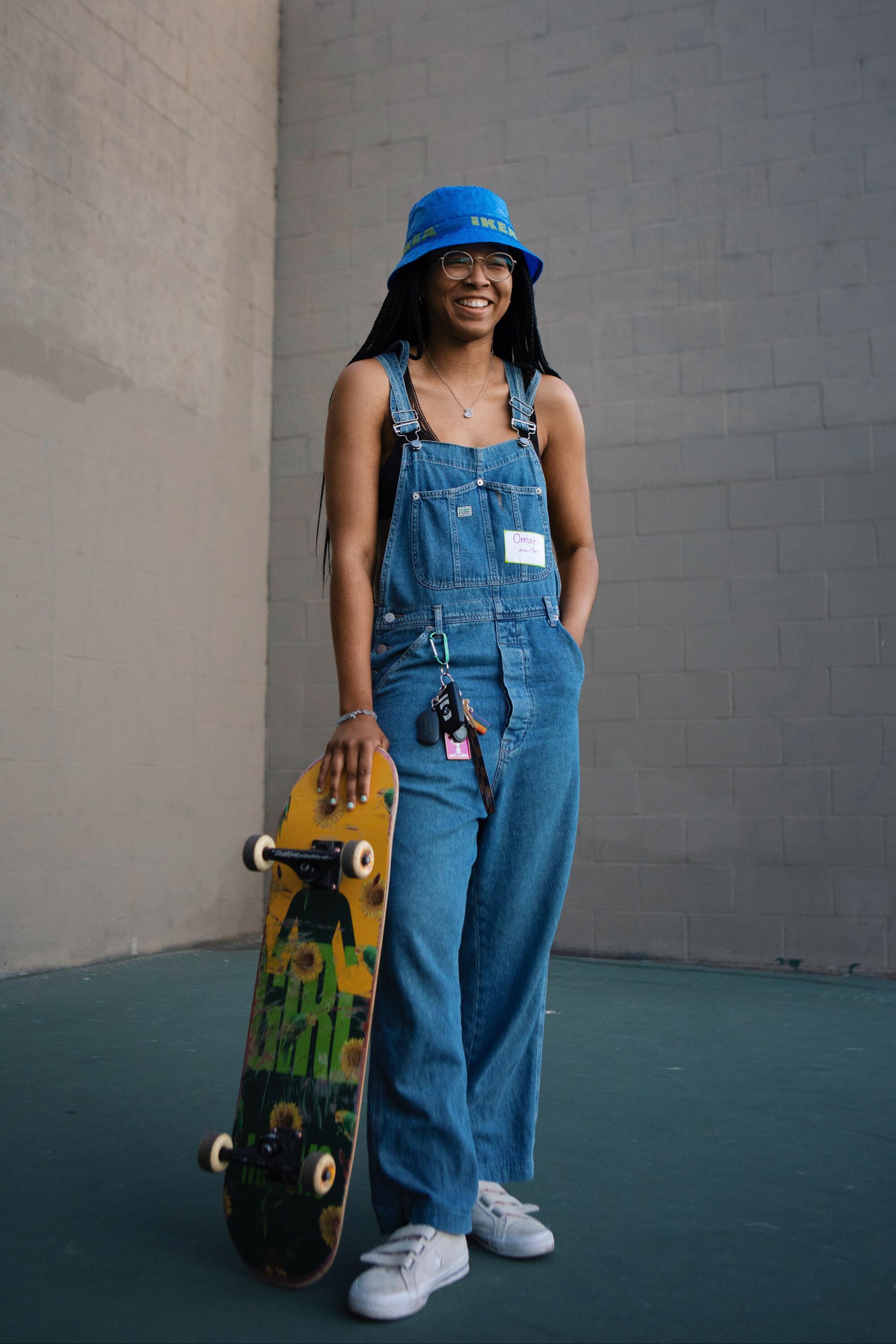 Omari Harmon poses for a portrait in denim overalls and blue bucket hat while holding a skateboard